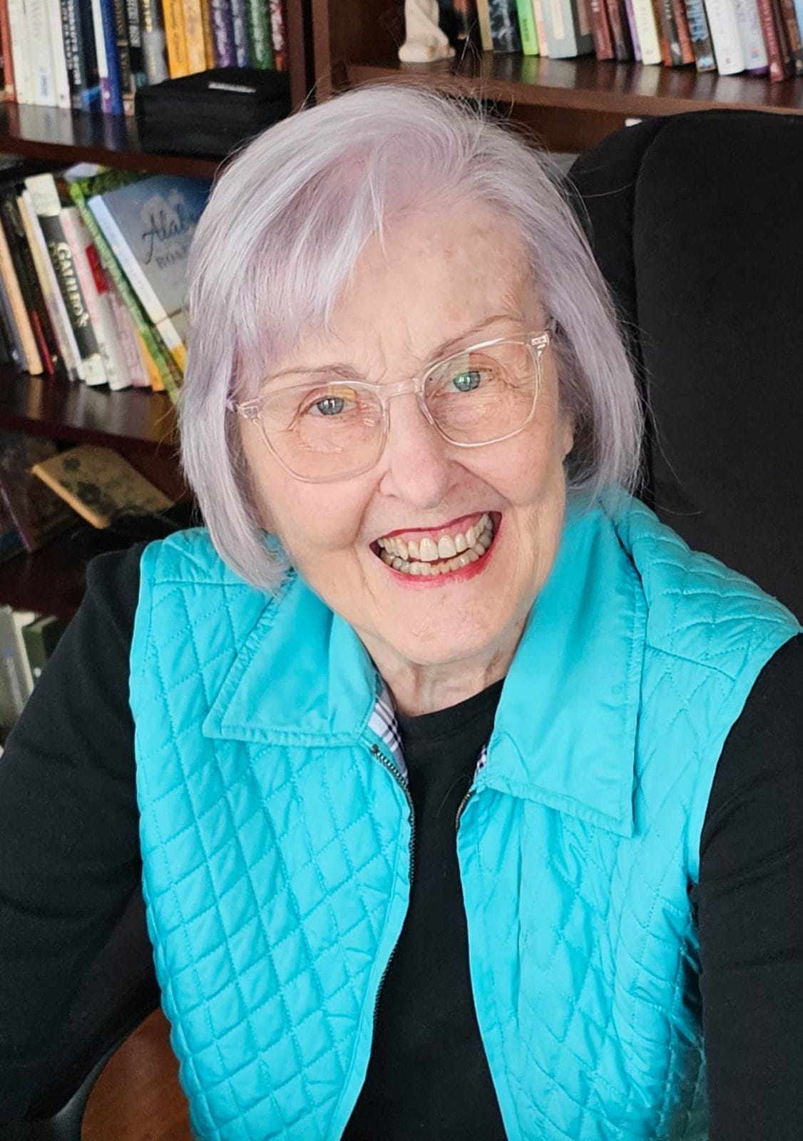 Elderly woman, Elizabeth Gunter Powell, with light purple hair wearing glasses and a blue vest, smiling in front of a bookshelf.
