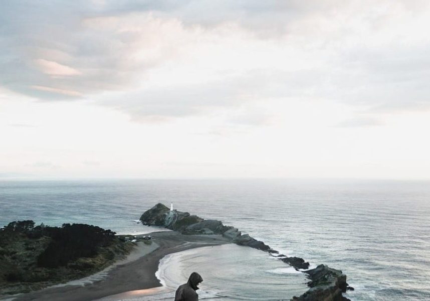 Person sitting on a cliff overlooking a scenic coastline at dusk.