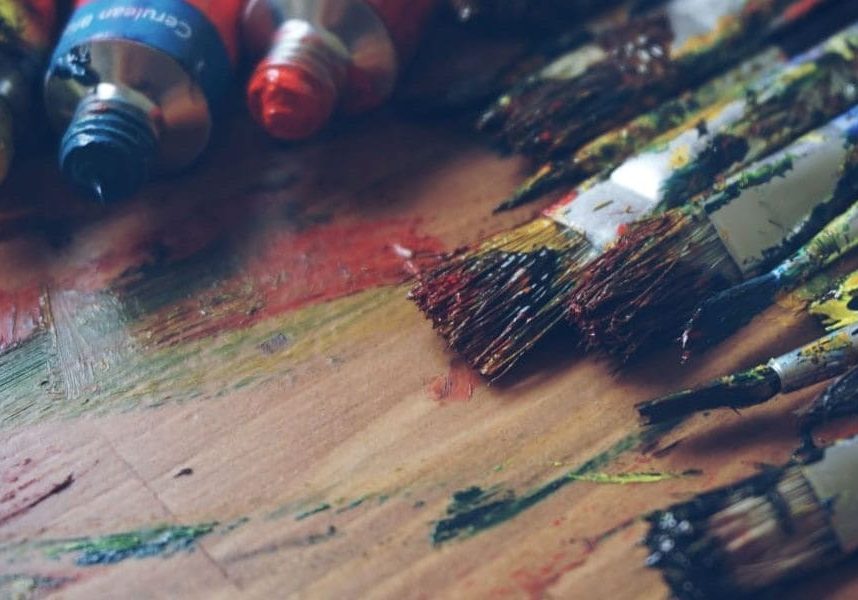 Assorted paintbrushes and tubes of paint on a stained artist's palette, tools in the crafting of God's Masterpiece.