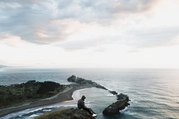 Person sitting on a cliff overlooking a scenic coastline at dusk.