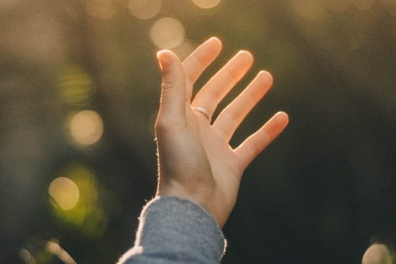 A human hand is extended towards a background of soft, golden light, with fingers slightly spread apart.