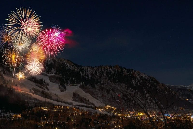 Fireworks illuminate the night sky above a snow-covered mountain slope with a lit village at its base, marking the blessing of 2024.